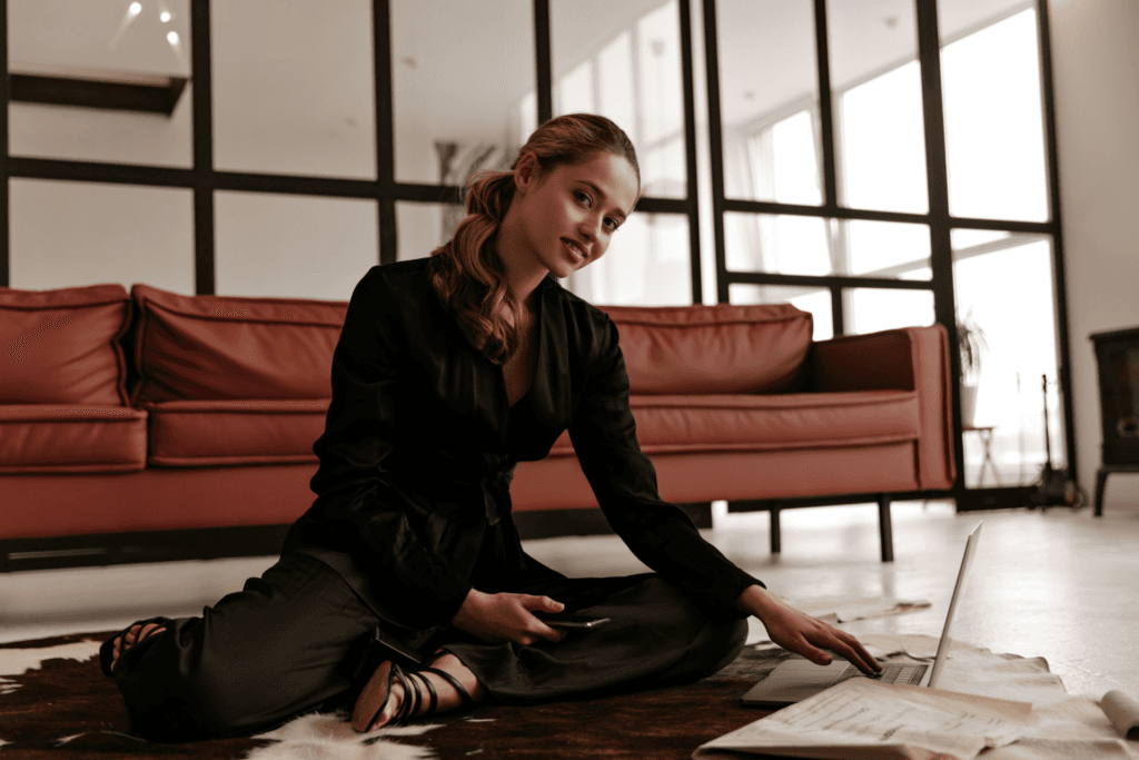 Confident woman in a black outfit working on a laptop while seated on the floor in a modern office, symbolizing strategic planning, entrepreneurship, and business growth