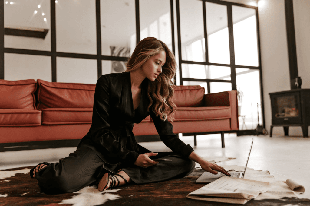 Confident woman in a black outfit working on a laptop while seated on the floor in a modern office, symbolizing strategic planning, entrepreneurship, and business growth