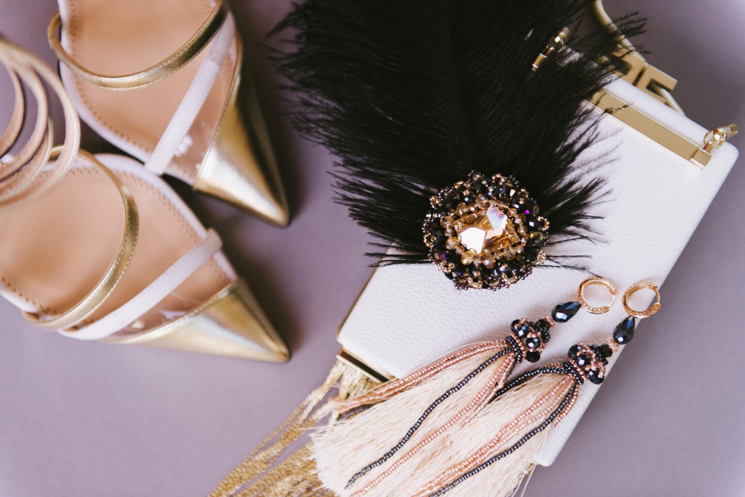 Flat lay of gold pointed-toe heels, a white clutch with feather embellishments, and beaded tassel earrings, symbolizing luxury fashion, premium accessories, and high-end retail branding.