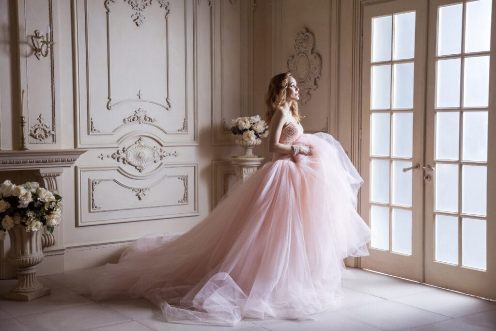 Elegant woman in a flowing blush pink ball gown standing in a luxurious, ornate room, symbolizing high-end fashion, bridal couture, and premium branding.