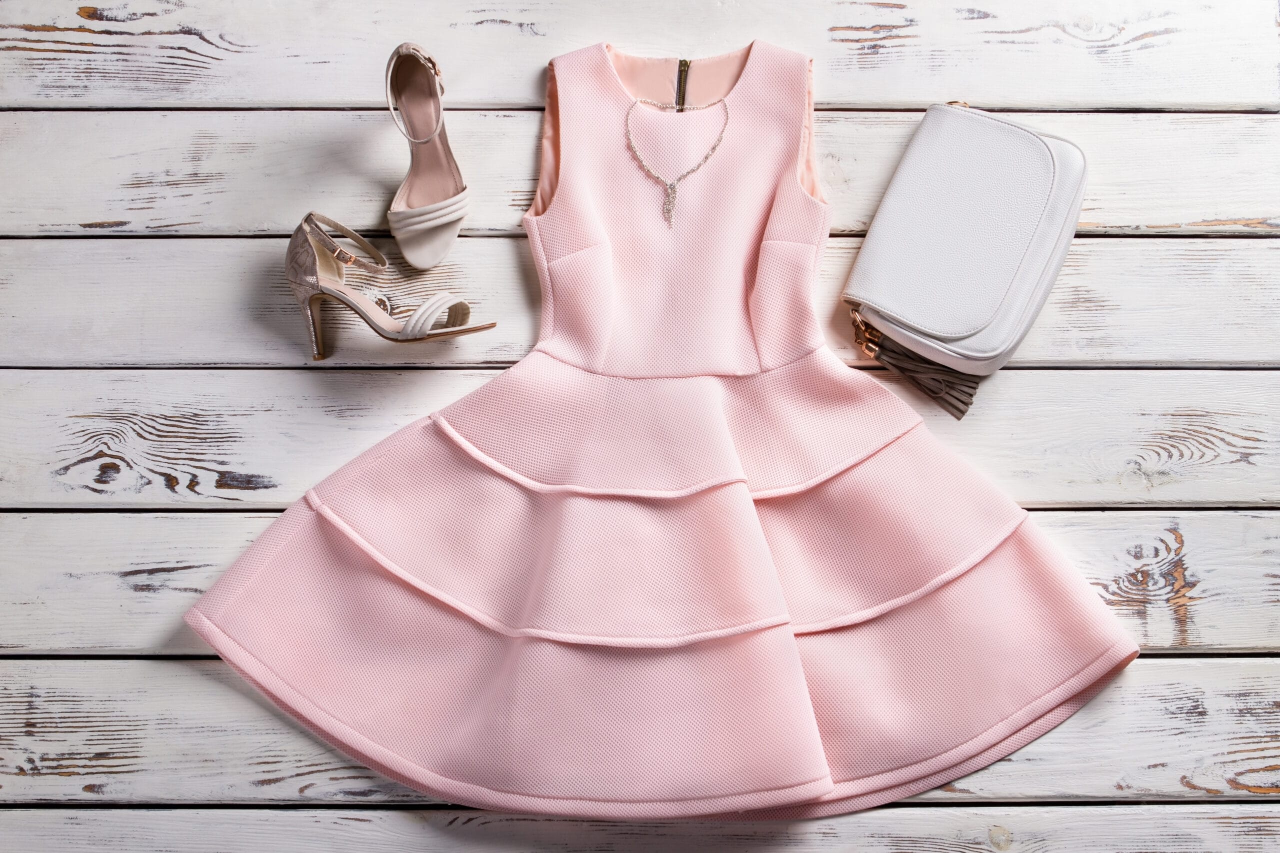 Flat lay of a pastel pink dress, matching heels, handbag, and accessories on a wooden background, representing fashion retail, merchandising, and brand styling.